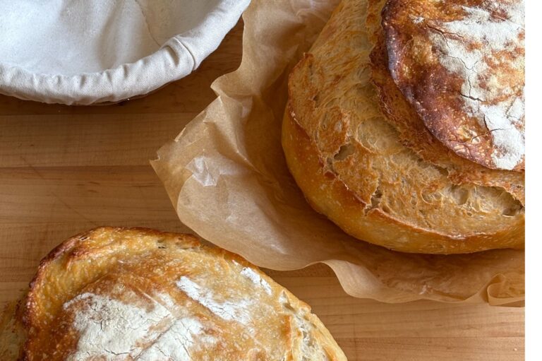 two loafs of sourdough on a surface. One is on a parchment paper and next to an empty sourdough banneton basket