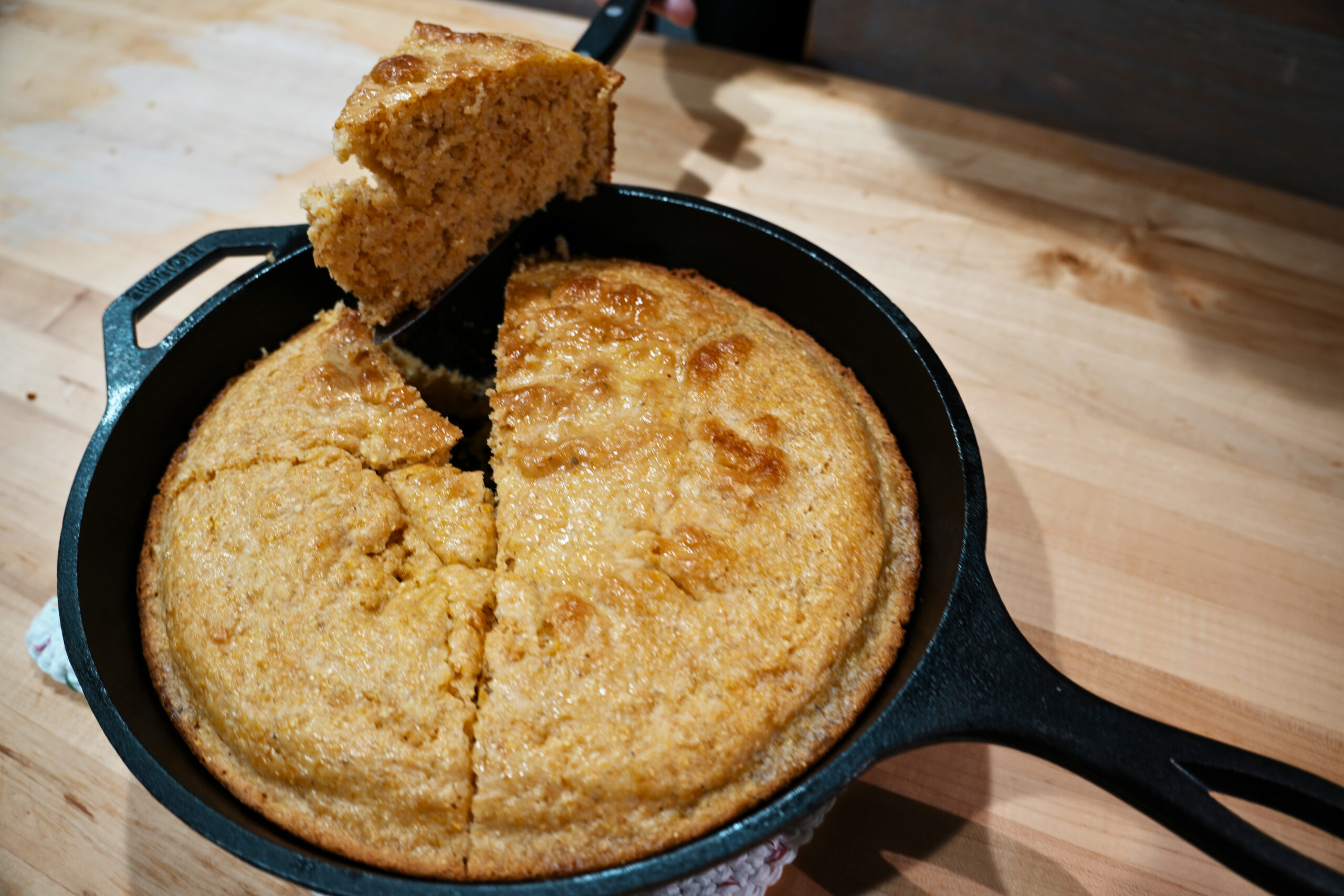 Cutting a slice of sourdough corn bread with that is fresh baked in a circular cast iron pan. The piece is cut as a triangle and resting on the edge of the pan